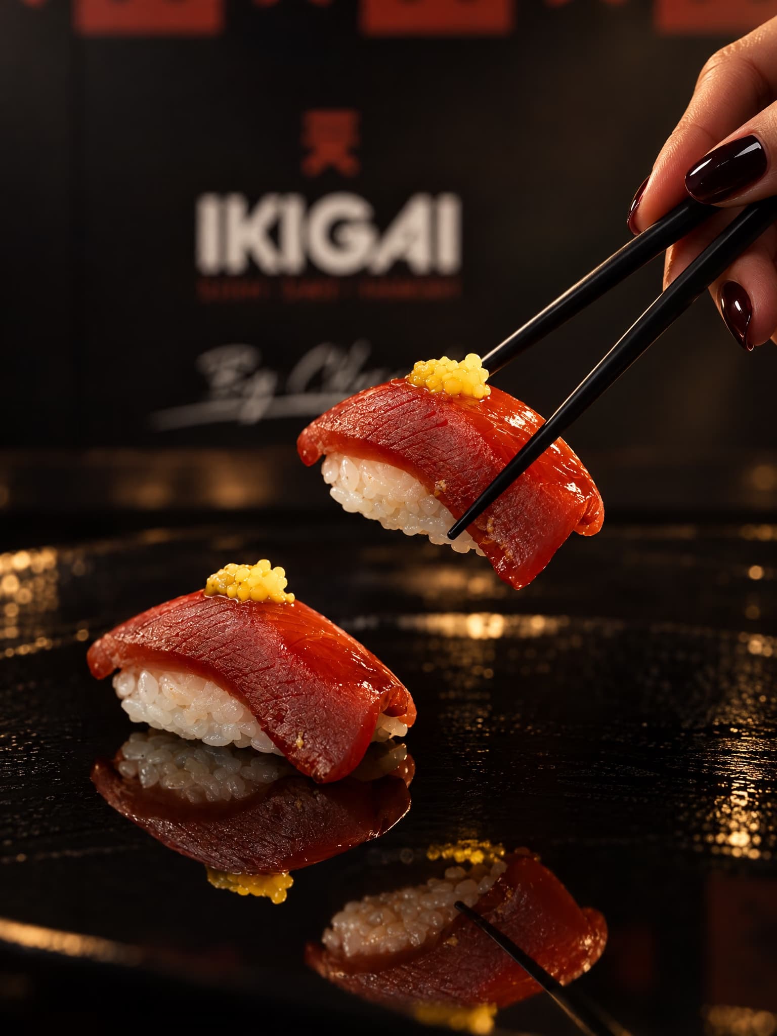 Unos palillos levantan un nigiri de atún con perlas amarillas, delante de una caja de Ikigai